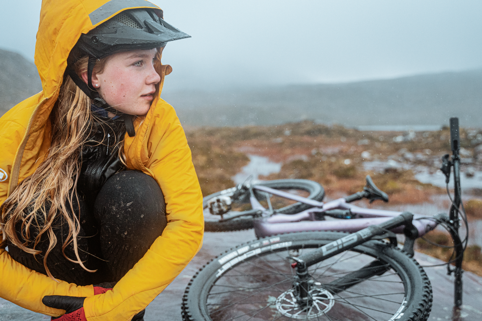 Kvinna i gul regnjacka sitter utomhus med lila terrängcykel